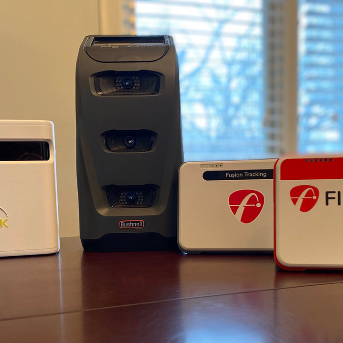 The SkyTrak+, Bushnell Launch Pro, FlightScope Mevo+ Limited Edition, and Mevo+ launch monitors standing together on a table with a window in the background