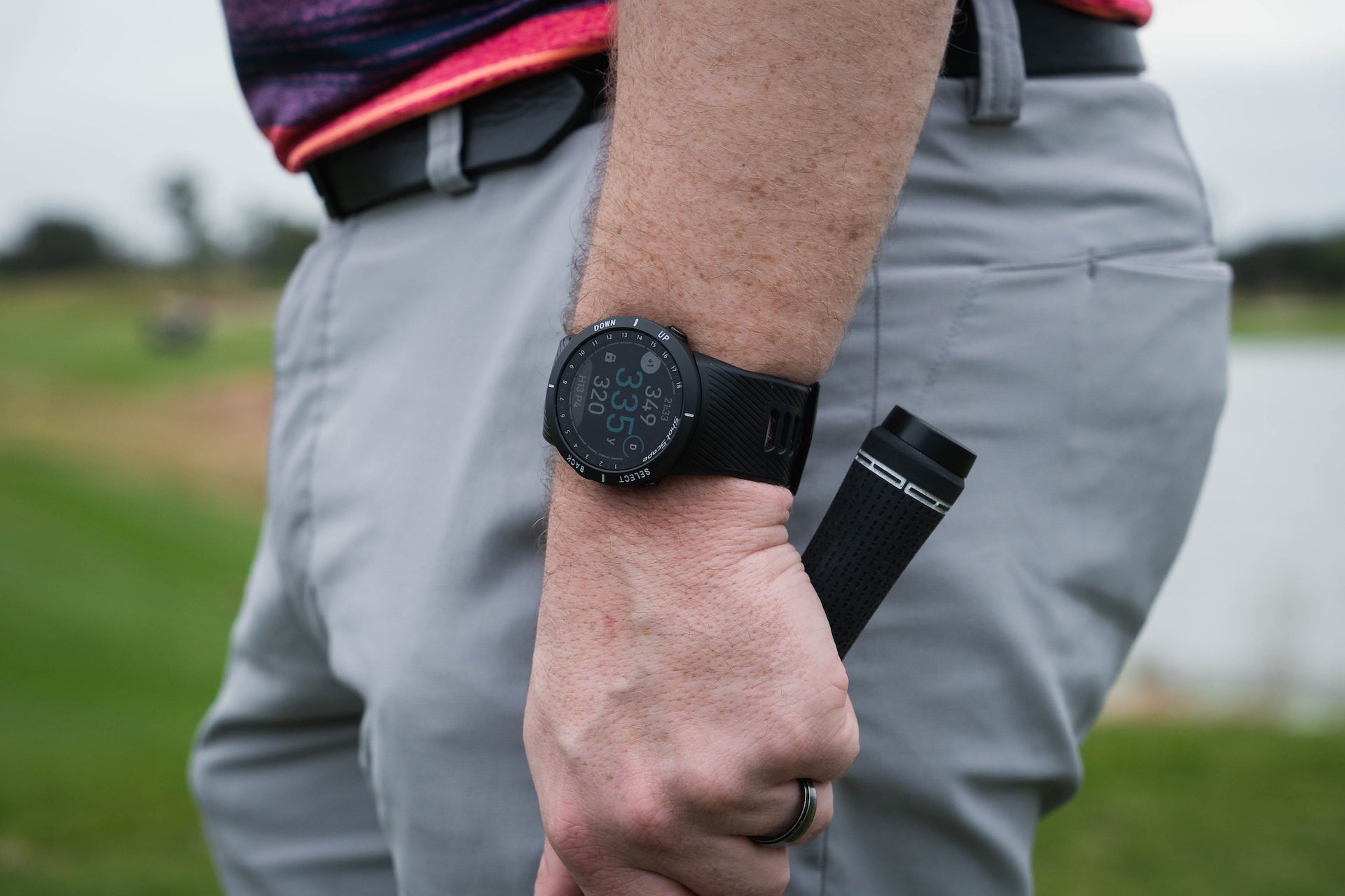 A Shot Scope V5 golf watch on a golfer's wrist while he's holding a golf club with a Shot Scope club tracker on it