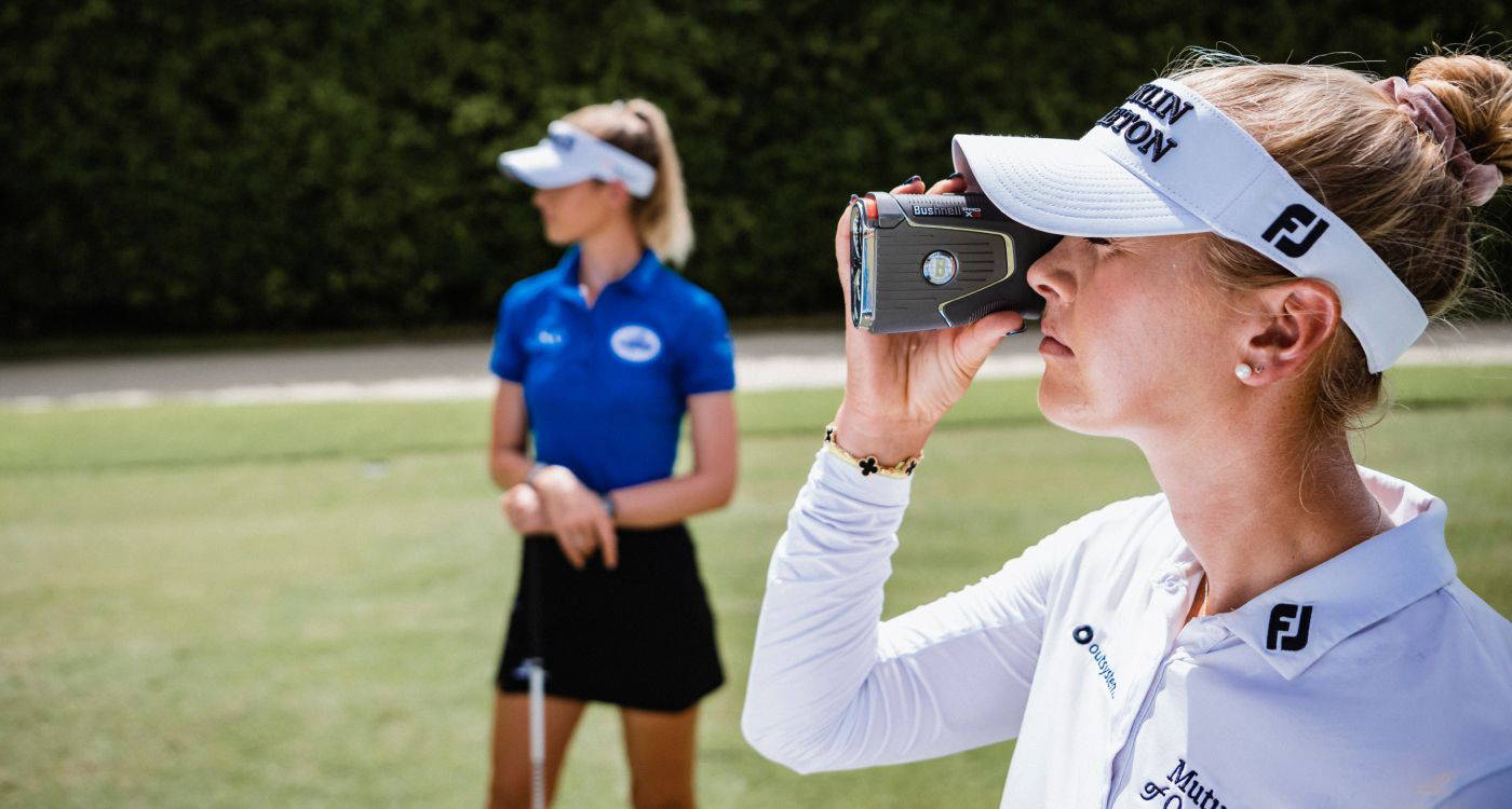 A golfer wearing a visor with a golf rangefinder to her face with another golfer in the background on the golf course