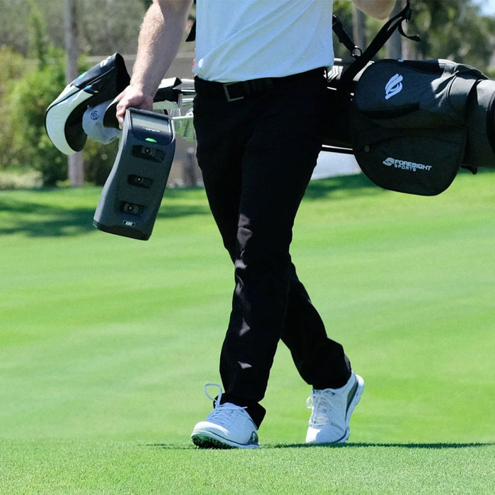 A golfer from the waist down walking on a golf course with a golf bag on his back and a Foresight Sports GC3 launch monitor in his hand
