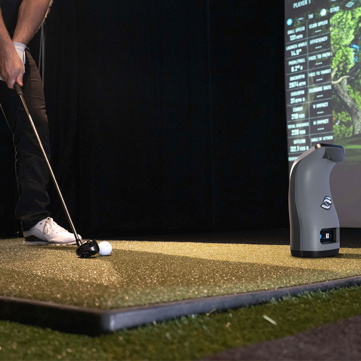 The lower part of a golfer with a club on a golf mat behind a golf ball and a Foresight Sports GC3 launch monitor all in an indoor golf simulator