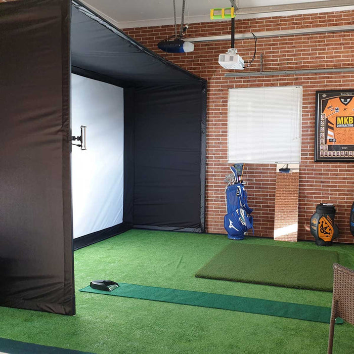 The PlayBetter SimStudio home golf simulator set up in a room with a brick wall and green turf on the floor with a ceiling mounted projector, golf hitting mat and putting green and blue golf bag leaning against the wall