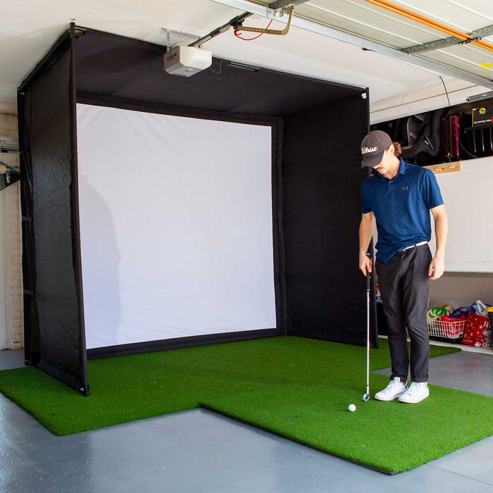 A golfer standing with a club at his side near a golf ball on a golf hitting mat in front of a PlayBetter SimStudio home golf simulator
